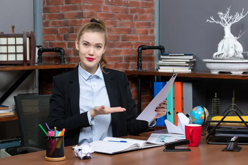 Assistante de direction assise à son bureau présentant un document, entourée de classeurs et fournitures de bureau