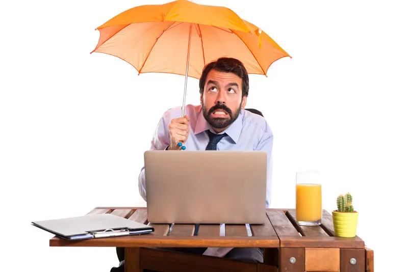Homme d'affaires à son bureau tenant un parapluie orange au-dessus de lui, illustrant la notion de protection et de couverture professionnelle
