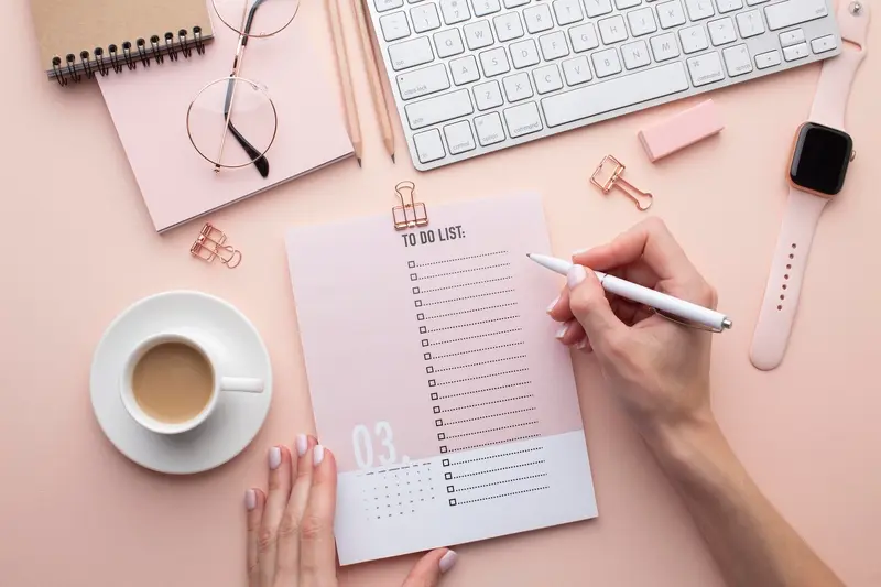 Main écrivant une liste de tâches sur un carnet "To Do List" entouré d'un clavier, d'une montre connectée et d'un café, sur un bureau rose