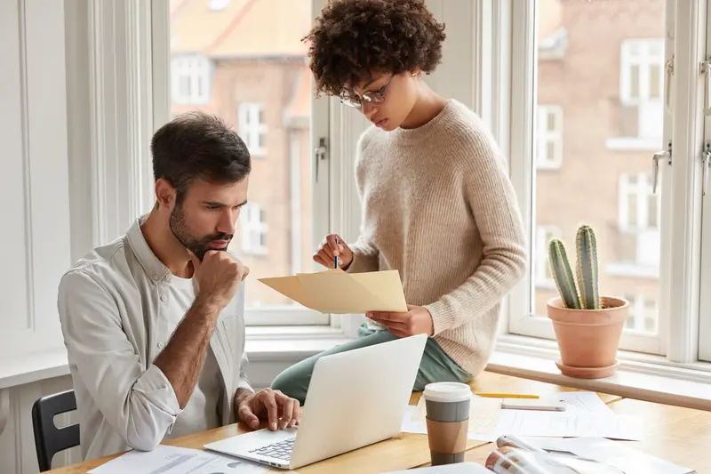 un jeune stagiaire assis sur un bureau à côté de son responsable, consultant des documents ce qui illustre l'accompagnement du stagiaire