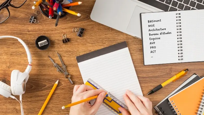 bureau avec bloc note, stylos, ordinateur portable et cahier où sont notés les termes techniques de la maîtrise d'oeuvre